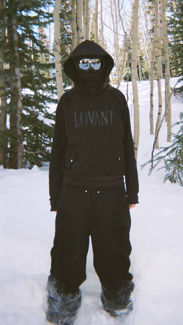 Model wearing the LOVANT black sweats and black LOVANT raglan hoodie with mirrored goggles and a face covering, standing in snow among aspen trees.