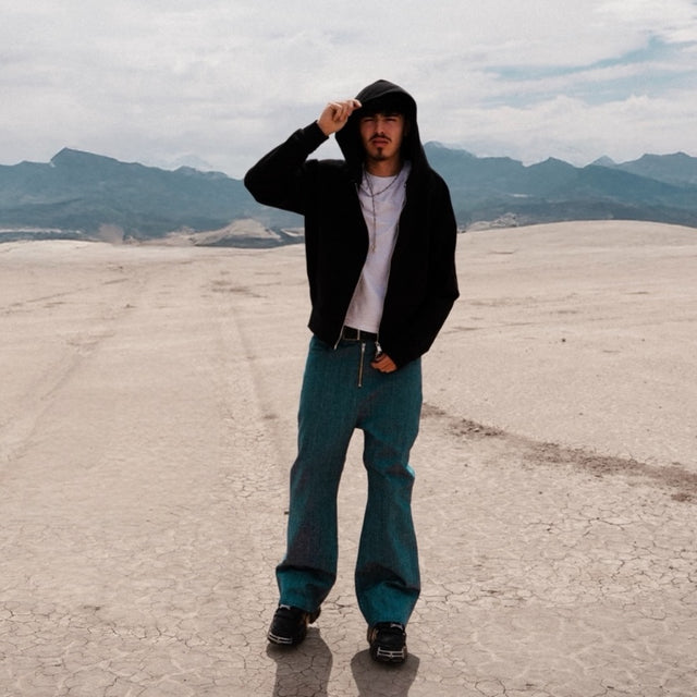 Model wearing the LOVANT blue UNIFORM flared denim with the black raglan UNIFORM zip-up hoodie and the white UNIFORM tee, standing on a cracked desert flat with mountains behind.